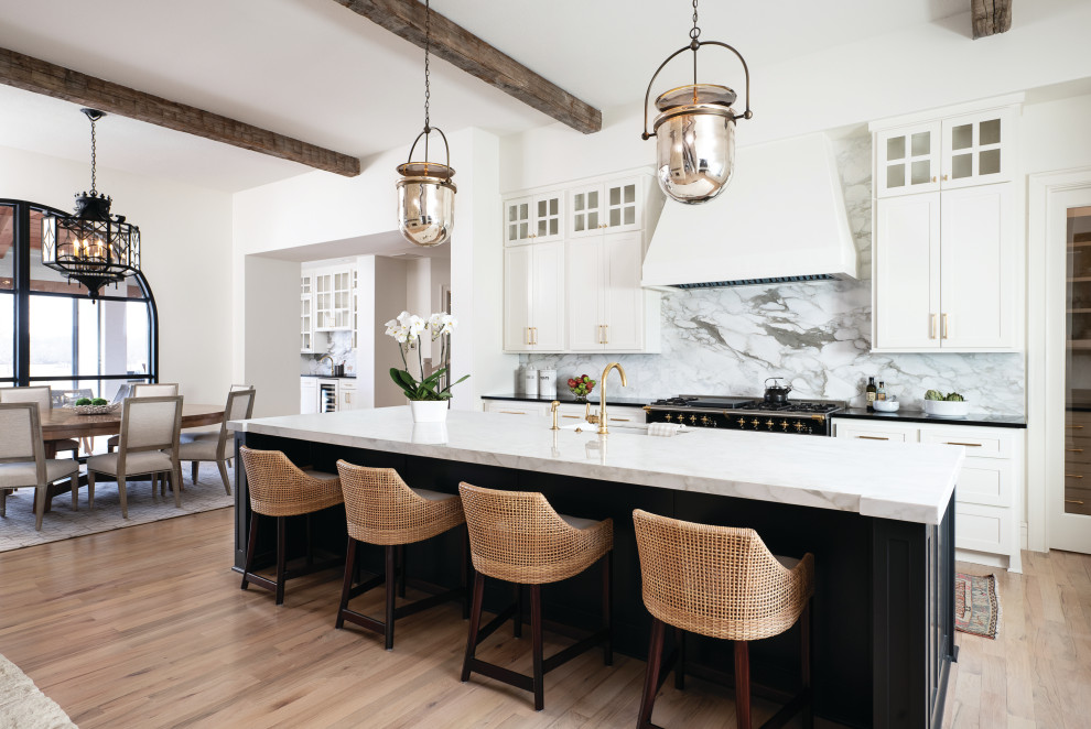This is an example of an expansive transitional galley open plan kitchen in Minneapolis with an undermount sink, recessed-panel cabinets, white cabinets, marble benchtops, white splashback, marble splashback, light hardwood floors, with island, white benchtop and exposed beam.