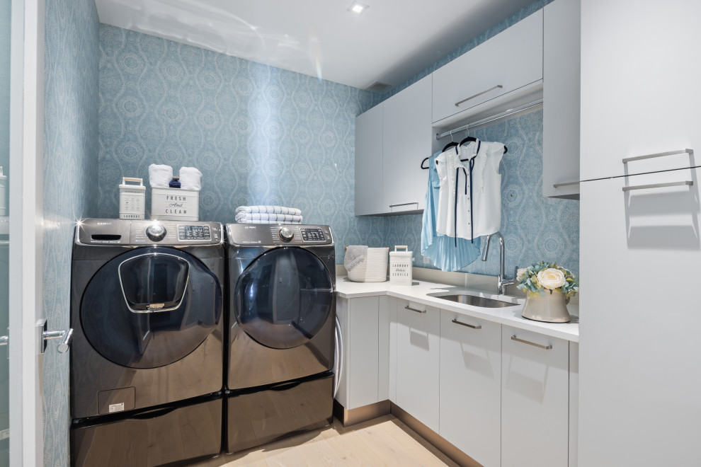 Idéer för att renovera en mellanstor vintage vita l-formad vitt tvättstuga enbart för tvätt, med en undermonterad diskho, släta luckor, grå skåp, bänkskiva i kvarts, vitt stänkskydd, blå väggar, ljust trägolv, en tvättmaskin och torktumlare bredvid varandra och beiget golv