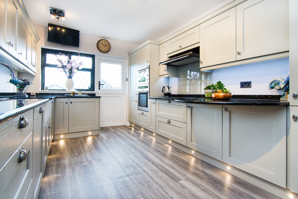 This is an example of a small traditional u-shaped separate kitchen in Essex with a farmhouse sink, shaker cabinets, light wood cabinets, granite benchtops, glass sheet splashback, black appliances, laminate floors, brown floor and multi-coloured benchtop.