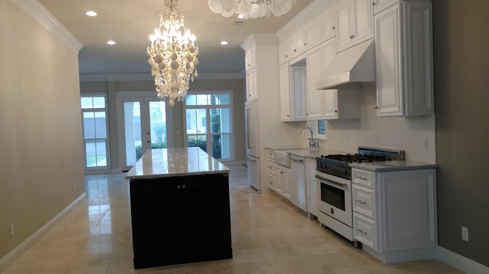 Photo of a mid-sized beach style single-wall eat-in kitchen in Miami with a farmhouse sink, raised-panel cabinets, white cabinets, marble benchtops, white splashback, ceramic splashback, white appliances, travertine floors and with island.