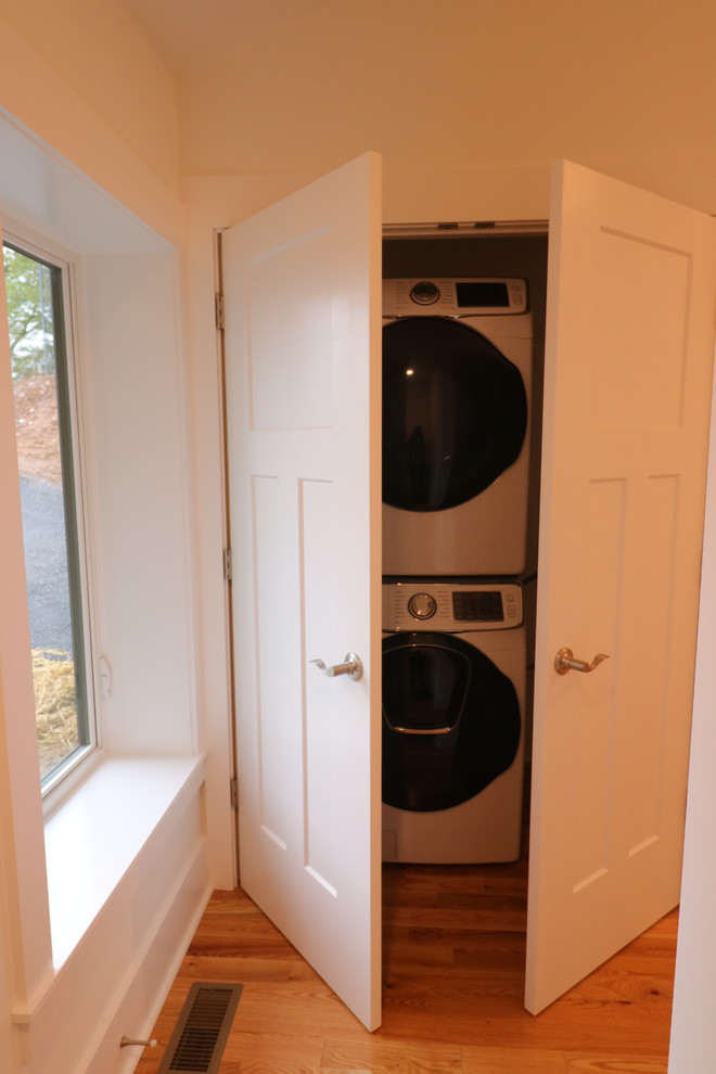 Inspiration for a small modern single-wall laundry cupboard in Other with white walls, light hardwood floors, a stacked washer and dryer and brown floor.