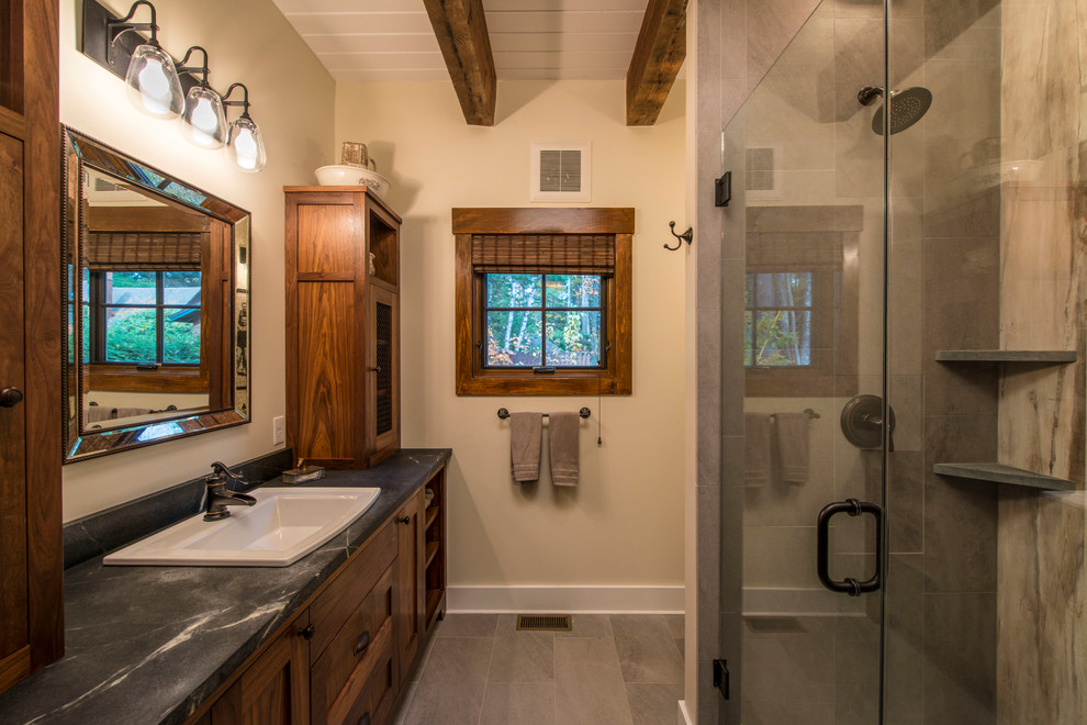 Photo of a small country bathroom in Boston with a drop-in sink, recessed-panel cabinets, medium wood cabinets, an open shower, gray tile, stone tile, beige walls, slate floors and soapstone benchtops.