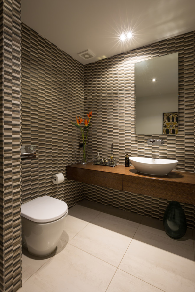 This is an example of a large contemporary powder room in Auckland with flat-panel cabinets, a one-piece toilet, multi-coloured tile, multi-coloured walls, porcelain floors, a vessel sink, wood benchtops, dark wood cabinets, brown benchtops, mosaic tile and beige floor.