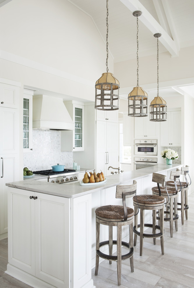 Inspiration for a large beach style l-shaped open plan kitchen in Atlanta with a farmhouse sink, recessed-panel cabinets, white cabinets, marble benchtops, stainless steel appliances, light hardwood floors, with island, white splashback, glass tile splashback and beige floor.