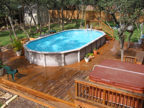  An abover-ground pool surrounded by a deck that is not raised to the level of the pool lip. Although this deck ties in the pool with a hot tub, and a few trees, for a landscaping element to this area