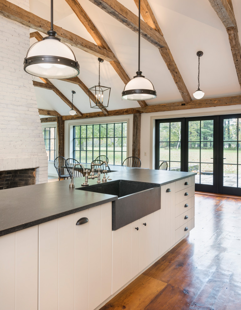 Photo of an expansive farmhouse u-shaped open plan kitchen in Boston with a belfast sink, beaded cabinets, white cabinets, black splashback, stainless steel appliances, medium hardwood flooring, an island, black worktops and exposed beams.