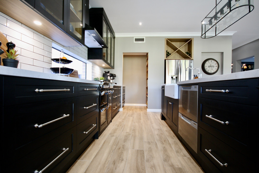 Large transitional single-wall open plan kitchen in Sydney with a farmhouse sink, shaker cabinets, black cabinets, quartz benchtops, white splashback, subway tile splashback, black appliances, light hardwood floors, with island, brown floor, white benchtop and coffered.