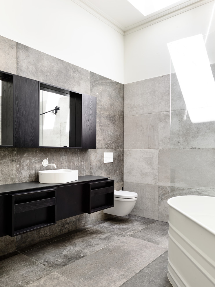 Inspiration for a mid-sized contemporary 3/4 bathroom in Melbourne with a freestanding tub, a wall-mount toilet, gray tile, ceramic tile, ceramic floors, grey floor, tile benchtops, black benchtops, flat-panel cabinets, black cabinets, white walls and a vessel sink.