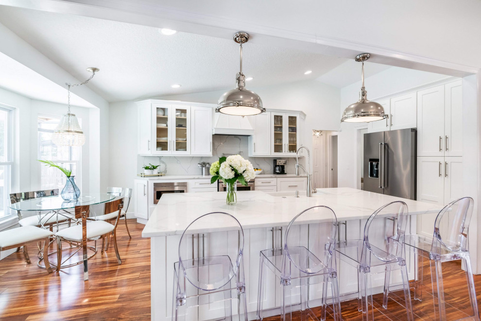 Photo of a large transitional u-shaped eat-in kitchen in Tampa with a farmhouse sink, shaker cabinets, white cabinets, quartzite benchtops, white splashback, engineered quartz splashback, stainless steel appliances, laminate floors, with island, brown floor, white benchtop and vaulted.