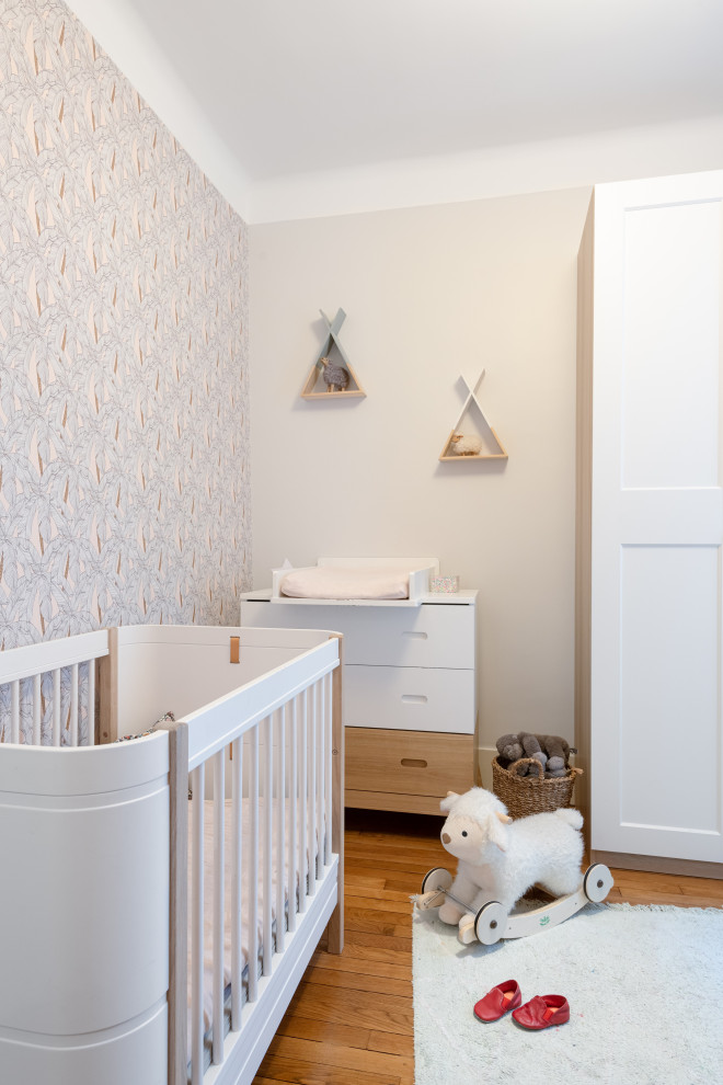 Imagen de habitación de bebé niña actual pequeña con paredes beige, suelo de madera clara y papel pintado
