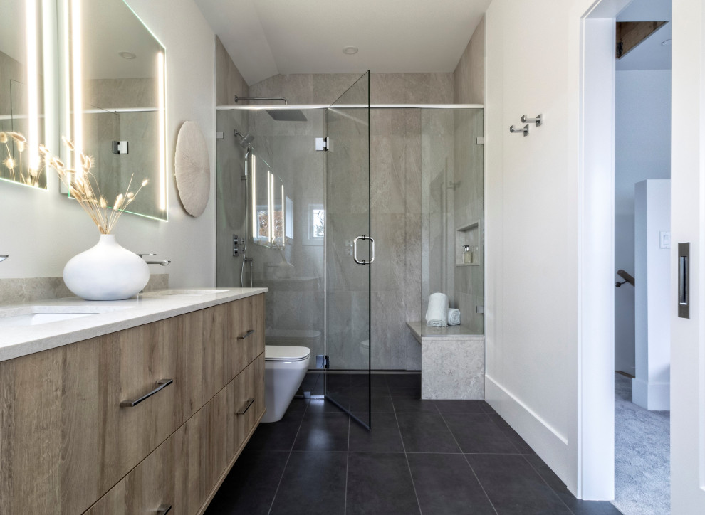 Design ideas for a large contemporary ensuite bathroom in Vancouver with flat-panel cabinets, medium wood cabinets, a freestanding bath, a built-in shower, a two-piece toilet, white tiles, stone slabs, white walls, porcelain flooring, a submerged sink, engineered stone worktops, black floors, a hinged door, white worktops, a shower bench, double sinks and a floating vanity unit.