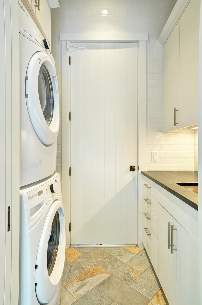 Inspiration for a mid-sized traditional galley dedicated laundry room in New York with an undermount sink, flat-panel cabinets, white cabinets, soapstone benchtops, white walls, travertine floors and a stacked washer and dryer.