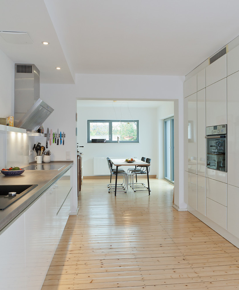 Inspiration for a mid-sized contemporary galley open plan kitchen in Cologne with flat-panel cabinets, white cabinets, light hardwood floors, no island, a drop-in sink, white splashback and stainless steel appliances.