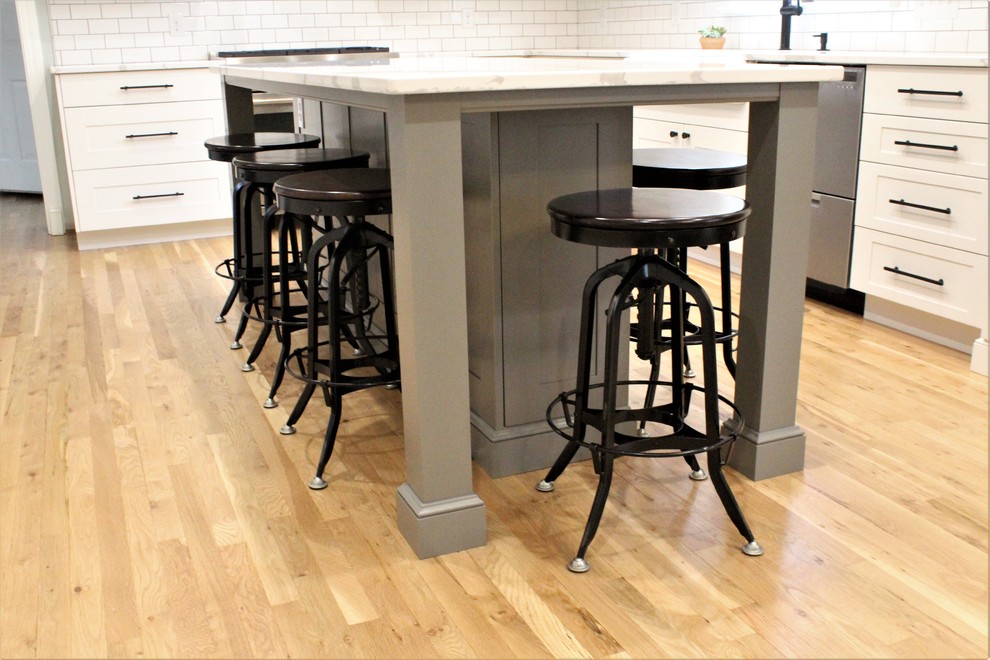 Example of a large transitional galley light wood floor eat-in kitchen design with a farmhouse sink, shaker cabinets, white cabinets, quartz countertops, white backsplash, ceramic backsplash, stainless steel appliances, an island and white countertops