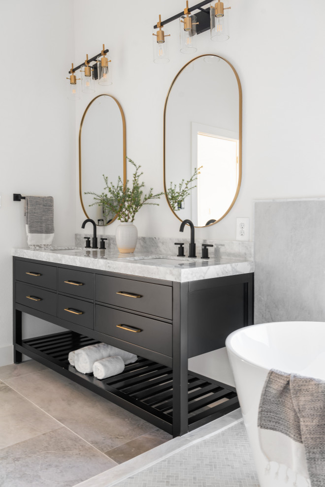This is an example of a mid-sized modern master bathroom in Dallas with recessed-panel cabinets, black cabinets, a freestanding tub, gray tile, porcelain tile, white walls, porcelain floors, an undermount sink, marble benchtops, grey floor, grey benchtops, an enclosed toilet, a double vanity and a freestanding vanity.