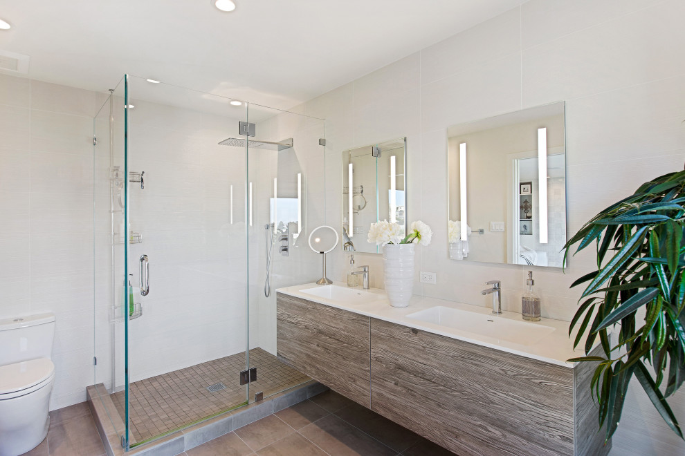 This is an example of a mid-sized modern master bathroom in San Francisco with flat-panel cabinets, medium wood cabinets, a corner shower, a one-piece toilet, white tile, porcelain tile, white walls, porcelain floors, an integrated sink, solid surface benchtops, brown floor, a hinged shower door, white benchtops, a double vanity and a floating vanity.