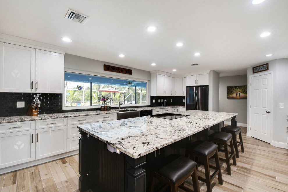 Inspiration for a large contemporary l-shaped eat-in kitchen in Miami with a farmhouse sink, shaker cabinets, white cabinets, granite benchtops, black splashback, glass tile splashback, stainless steel appliances, ceramic floors, with island, grey floor and multi-coloured benchtop.
