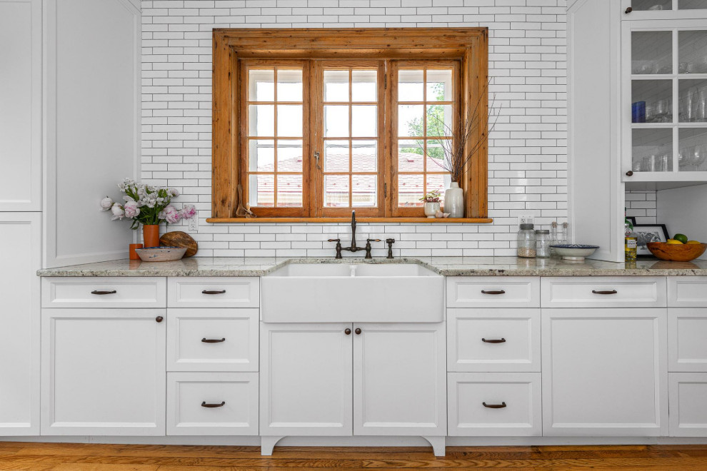 Example of a small transitional galley light wood floor and brown floor enclosed kitchen design in Ottawa with a farmhouse sink, shaker cabinets, white cabinets, granite countertops, white backsplash, subway tile backsplash, stainless steel appliances, an island and gray countertops