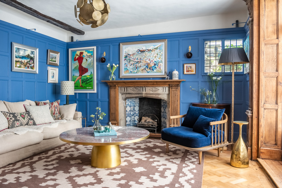 Large eclectic enclosed living room in London with a library, blue walls, light hardwood floors, a standard fireplace, a stone fireplace surround, a concealed tv, beige floor, exposed beam and wood walls.