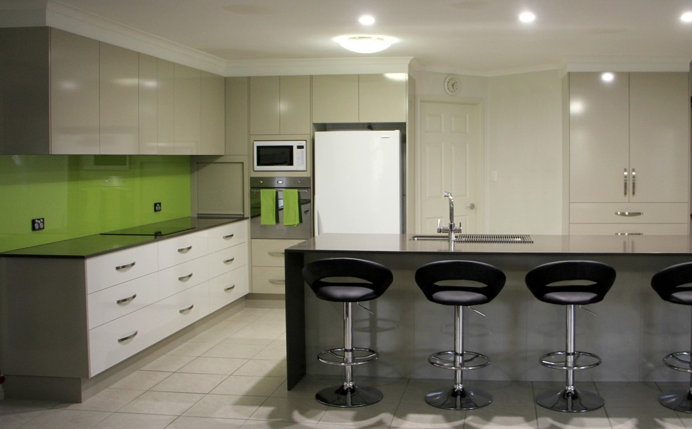 Design ideas for a large contemporary l-shaped kitchen in Brisbane with a double-bowl sink, flat-panel cabinets, beige cabinets, granite benchtops, green splashback, glass sheet splashback, white appliances, porcelain floors and with island.