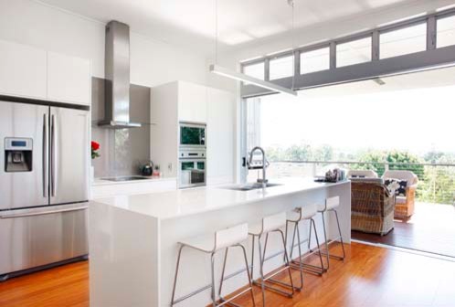 This is an example of a mid-sized transitional single-wall open plan kitchen in Brisbane with an undermount sink, flat-panel cabinets, white cabinets, solid surface benchtops, grey splashback, glass sheet splashback, stainless steel appliances, medium hardwood floors and with island.