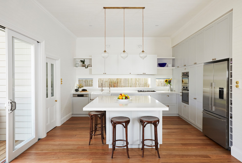 Mid-sized transitional l-shaped open plan kitchen in Sydney with a farmhouse sink, shaker cabinets, grey cabinets, solid surface benchtops, white splashback, window splashback, stainless steel appliances, with island and white benchtop.