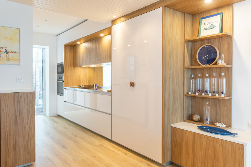 Bild på ett litet maritimt kök, med en integrerad diskho, luckor med profilerade fronter, vita skåp, bänkskiva i rostfritt stål, spegel som stänkskydd, rostfria vitvaror och ljust trägolv