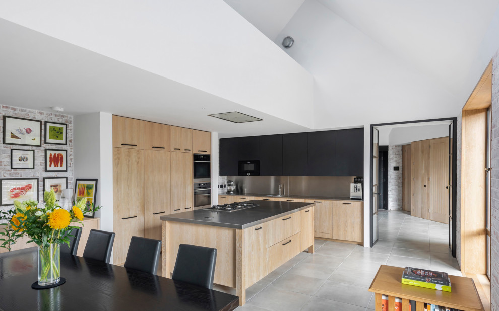 Photo of a large contemporary l-shaped eat-in kitchen in Sussex with a double-bowl sink, flat-panel cabinets, light wood cabinets, stainless steel benchtops, grey splashback, black appliances, ceramic floors, with island, grey floor and grey benchtop.