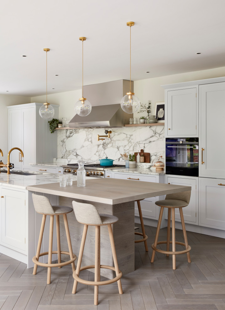 Photo of a large transitional galley open plan kitchen in Hampshire with a drop-in sink, shaker cabinets, grey cabinets, marble benchtops, white splashback, marble splashback, stainless steel appliances, light hardwood floors, with island, beige floor and white benchtop.