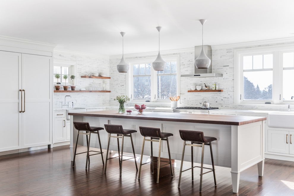 This is an example of a transitional l-shaped kitchen in Portland Maine with a farmhouse sink, shaker cabinets, white cabinets, multi-coloured splashback, panelled appliances, dark hardwood floors, with island, brown floor, white benchtop and marble splashback.