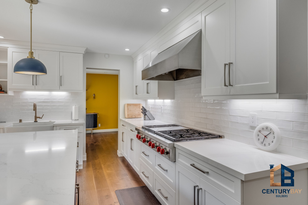 Elegant l-shaped light wood floor and brown floor kitchen photo in San Francisco with a farmhouse sink, raised-panel cabinets, white cabinets, quartz countertops, white backsplash, porcelain backsplash, stainless steel appliances, an island and multicolored countertops