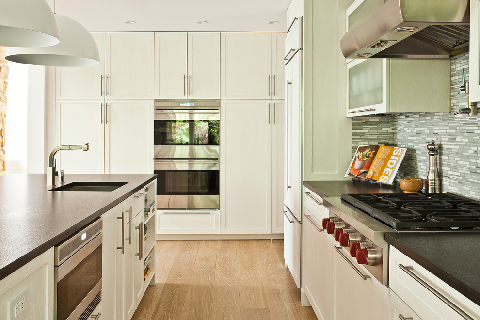 Photo of a large contemporary u-shaped eat-in kitchen in New York with an undermount sink, glass-front cabinets, white cabinets, grey splashback, stainless steel appliances, light hardwood floors and with island.