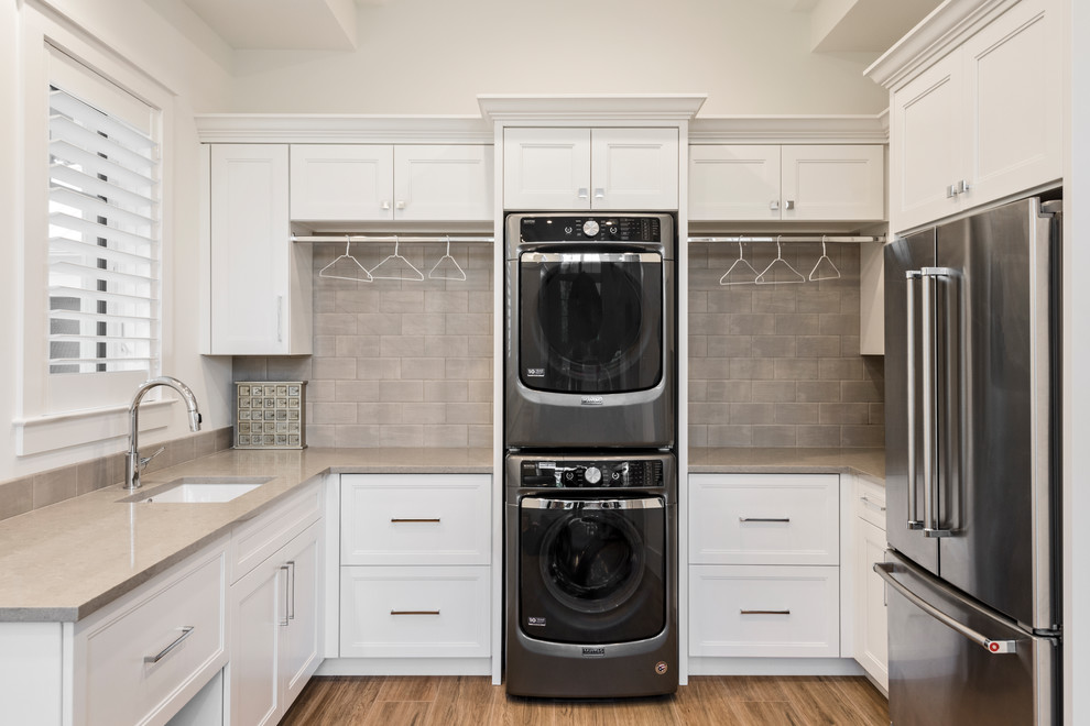 This is an example of an expansive transitional laundry room in Miami with a drop-in sink, recessed-panel cabinets, white cabinets, granite benchtops, white walls, medium hardwood floors and a stacked washer and dryer.
