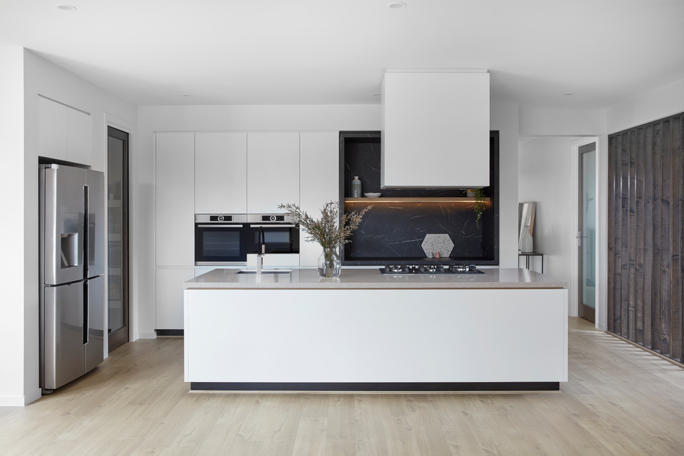 This is an example of a contemporary kitchen in Melbourne with an undermount sink, flat-panel cabinets, white cabinets, black splashback, stone slab splashback, stainless steel appliances, light hardwood floors, with island, beige floor and black benchtop.