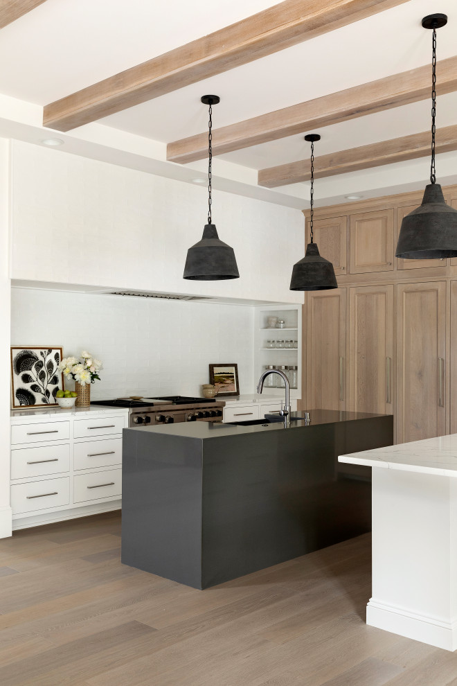 Design ideas for a beach style l-shaped kitchen in Minneapolis with a farmhouse sink, flat-panel cabinets, white cabinets, white splashback, stainless steel appliances, light hardwood floors, multiple islands, white benchtop, exposed beam and brown floor.
