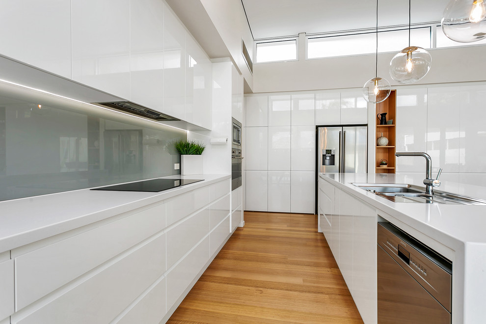 Contemporary u-shaped kitchen in Adelaide with flat-panel cabinets, white cabinets, grey splashback, glass sheet splashback, stainless steel appliances, medium hardwood floors, with island, brown floor and white benchtop.