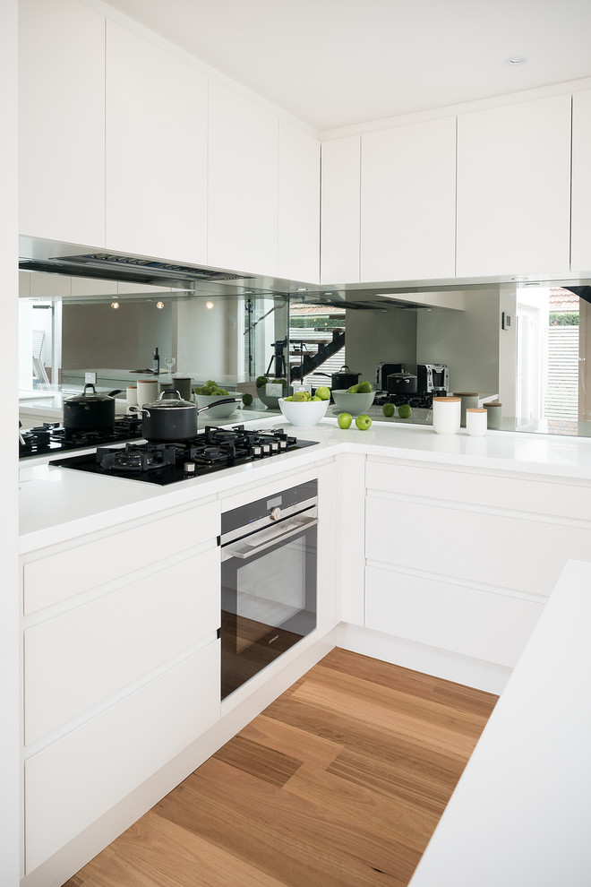 This is an example of a mid-sized contemporary l-shaped eat-in kitchen in Melbourne with an integrated sink, flat-panel cabinets, white cabinets, solid surface benchtops, mirror splashback, stainless steel appliances, medium hardwood floors, with island, metallic splashback and white benchtop.