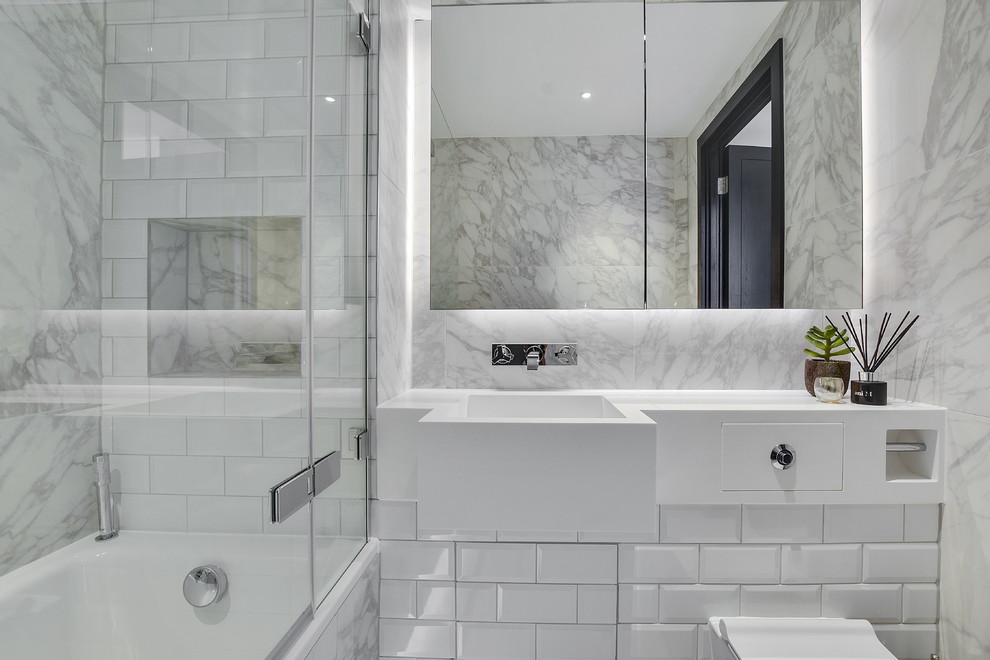 This is an example of a small contemporary kids bathroom in London with glass-front cabinets, white cabinets, a drop-in tub, a shower/bathtub combo, a wall-mount toilet, white tile, marble, white walls, porcelain floors, an integrated sink, engineered quartz benchtops, white floor and a hinged shower door.