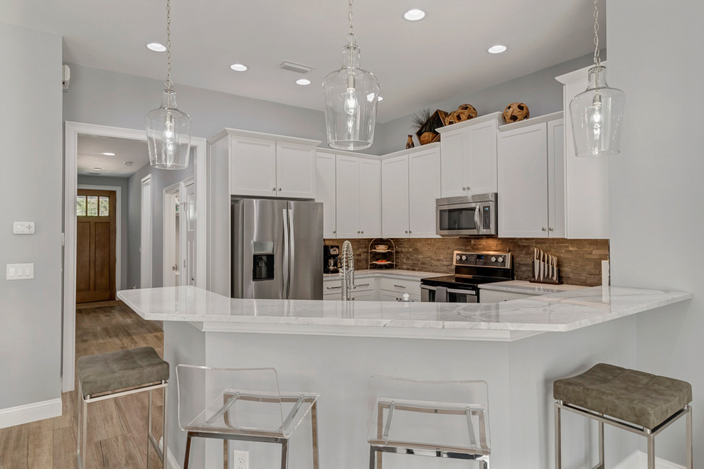 Mid-sized beach style u-shaped eat-in kitchen in Jacksonville with a farmhouse sink, shaker cabinets, white cabinets, quartzite benchtops, slate splashback, stainless steel appliances, light hardwood floors, with island, brown floor, white benchtop and grey splashback.