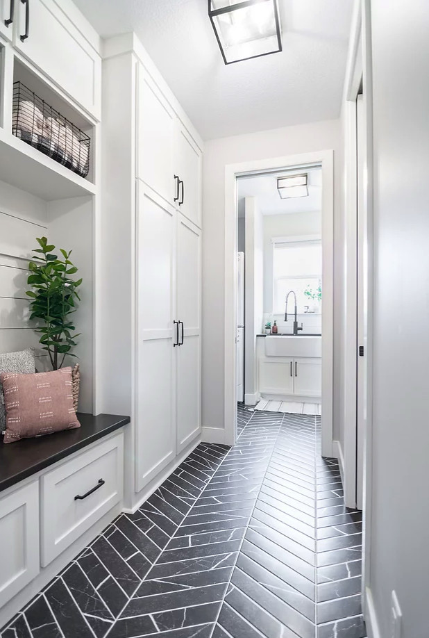 Photo of a mid-sized transitional galley utility room in Minneapolis with a farmhouse sink, shaker cabinets, white cabinets, quartz benchtops, white splashback, ceramic splashback, grey walls, ceramic floors, a stacked washer and dryer, black floor and black benchtop.