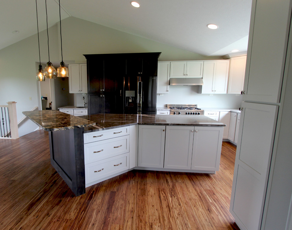 Large transitional l-shaped kitchen in Cleveland with a farmhouse sink, flat-panel cabinets, white cabinets, granite benchtops, white splashback, ceramic splashback, stainless steel appliances, medium hardwood floors, with island, brown floor and multi-coloured benchtop.