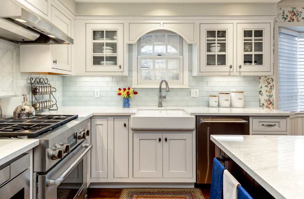 Photo of a country kitchen/diner in Denver with a belfast sink, glass-front cabinets, white cabinets, composite countertops, white splashback, metro tiled splashback, stainless steel appliances, medium hardwood flooring, an island and white worktops.