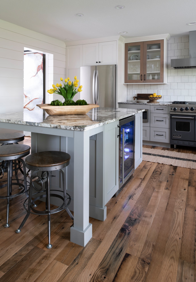 This is an example of a mid-sized country l-shaped eat-in kitchen in Minneapolis with a farmhouse sink, shaker cabinets, white cabinets, white splashback, ceramic splashback, stainless steel appliances, medium hardwood floors, with island, brown floor and multi-coloured benchtop.