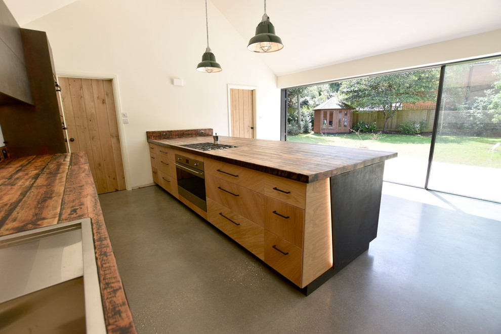 This is an example of a large industrial single-wall open plan kitchen in Oxfordshire with a double-bowl sink, flat-panel cabinets, light wood cabinets, wood benchtops, white splashback, glass sheet splashback, black appliances, concrete floors, with island, grey floor and multi-coloured benchtop.