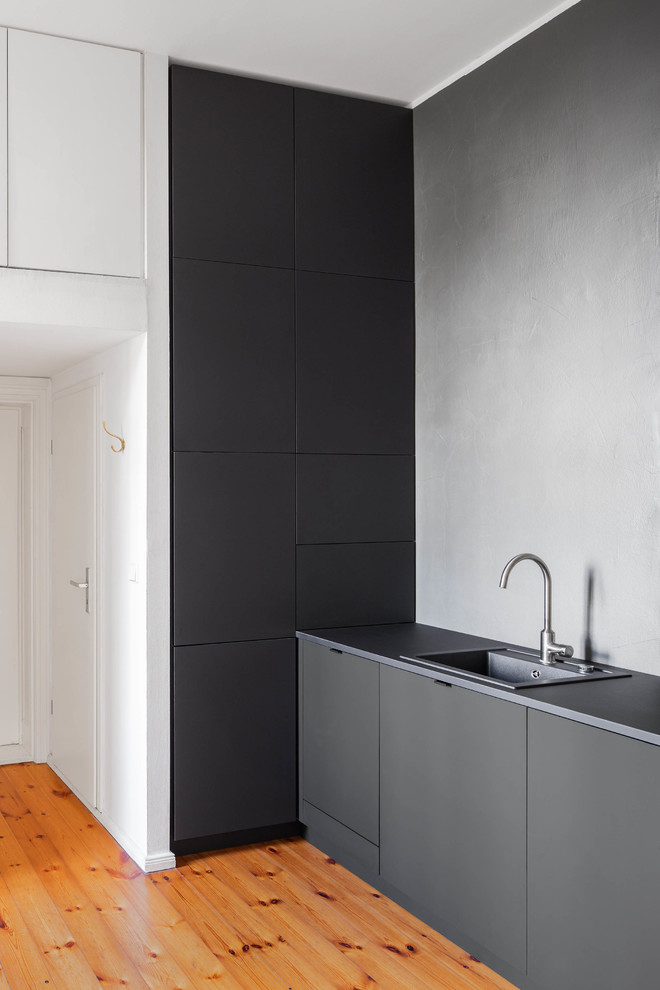 This is an example of a mid-sized modern single-wall open plan kitchen in Berlin with a drop-in sink, flat-panel cabinets, black cabinets, solid surface benchtops, black splashback, stone slab splashback, stainless steel appliances, medium hardwood floors, no island, brown floor and black benchtop.