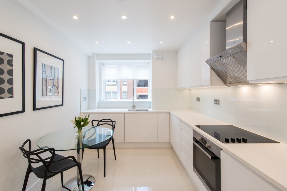 This is an example of a contemporary l-shaped eat-in kitchen in London with a double-bowl sink, flat-panel cabinets, white cabinets, white splashback, glass sheet splashback, black appliances and no island.