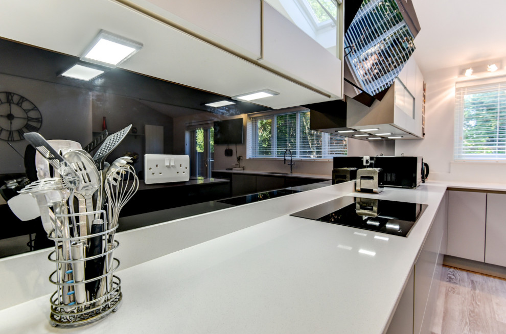 Large modern grey and white l-shaped kitchen/diner in Sussex with a submerged sink, flat-panel cabinets, grey cabinets, quartz worktops, black splashback, glass sheet splashback, black appliances, vinyl flooring, an island, grey floors, white worktops and feature lighting.