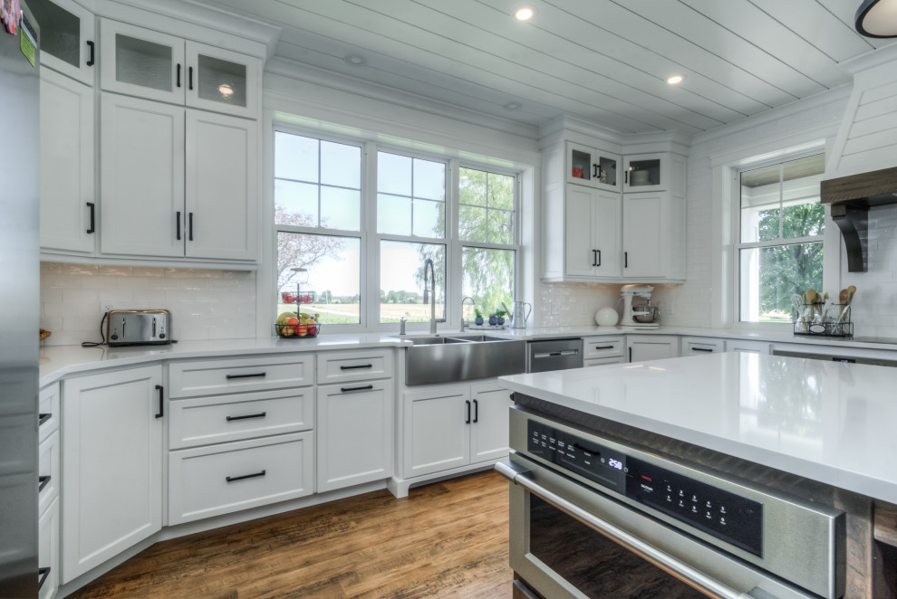 Large country u-shaped kitchen in Toronto with a farmhouse sink, shaker cabinets, white cabinets, quartz benchtops, white splashback, cement tile splashback, stainless steel appliances, medium hardwood floors, with island, multi-coloured floor, white benchtop and timber.