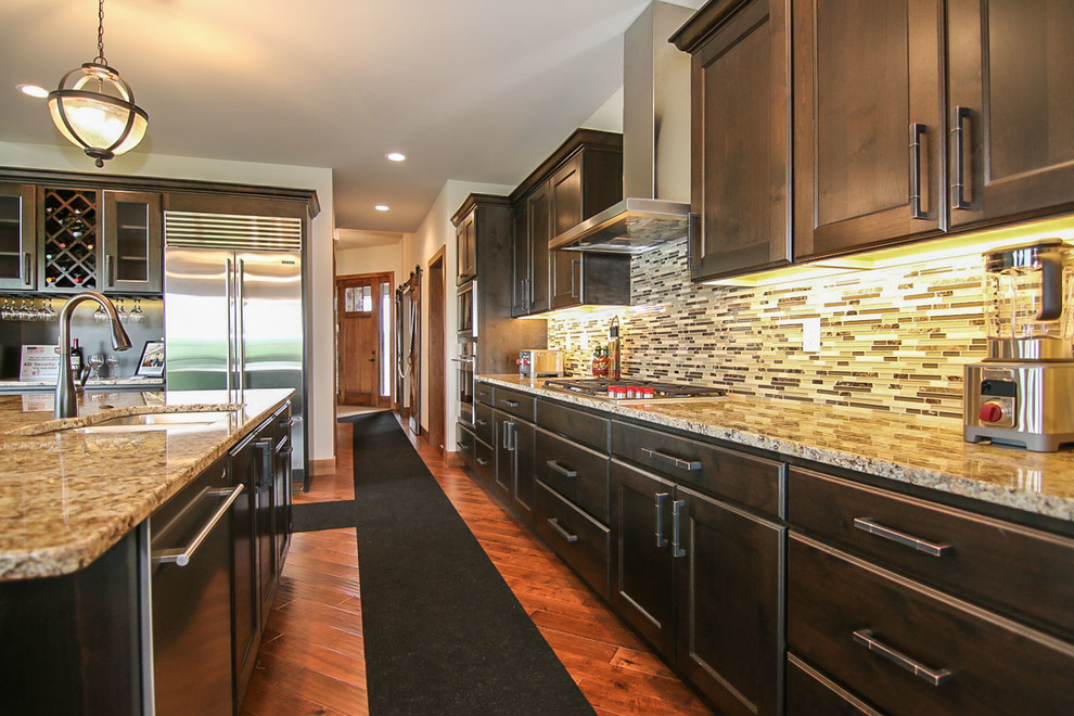 Example of a large transitional medium tone wood floor and brown floor kitchen design with an undermount sink, recessed-panel cabinets, dark wood cabinets, granite countertops, multicolored backsplash, glass tile backsplash, stainless steel appliances and an island