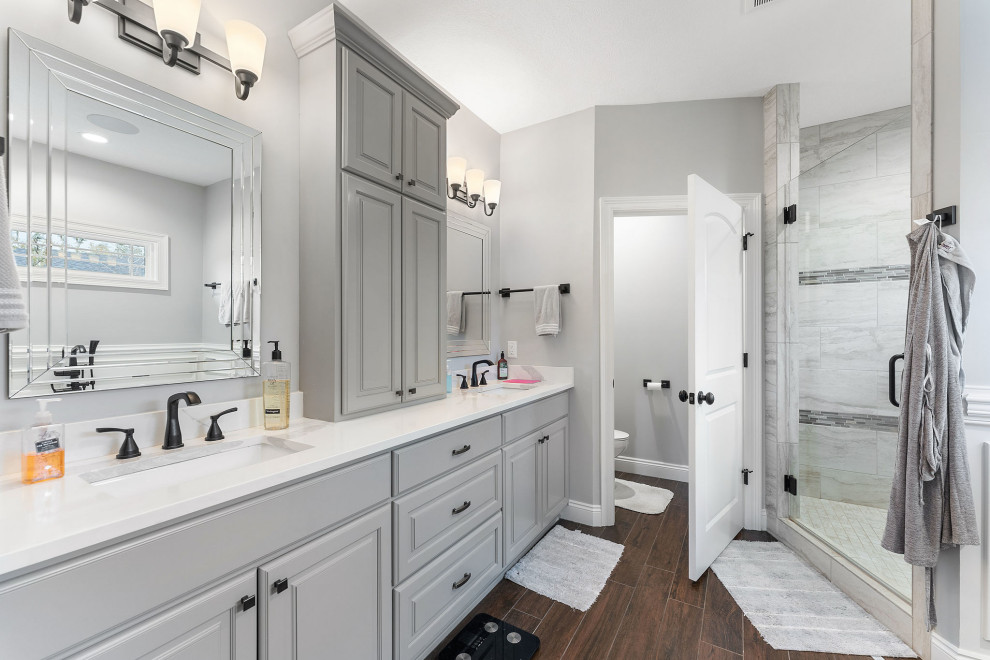 This is an example of a mid-sized traditional master bathroom in Other with raised-panel cabinets, grey cabinets, a freestanding tub, a double shower, a two-piece toilet, gray tile, porcelain tile, grey walls, porcelain floors, an undermount sink, engineered quartz benchtops, brown floor, a hinged shower door, white benchtops, an enclosed toilet, a double vanity and a built-in vanity.